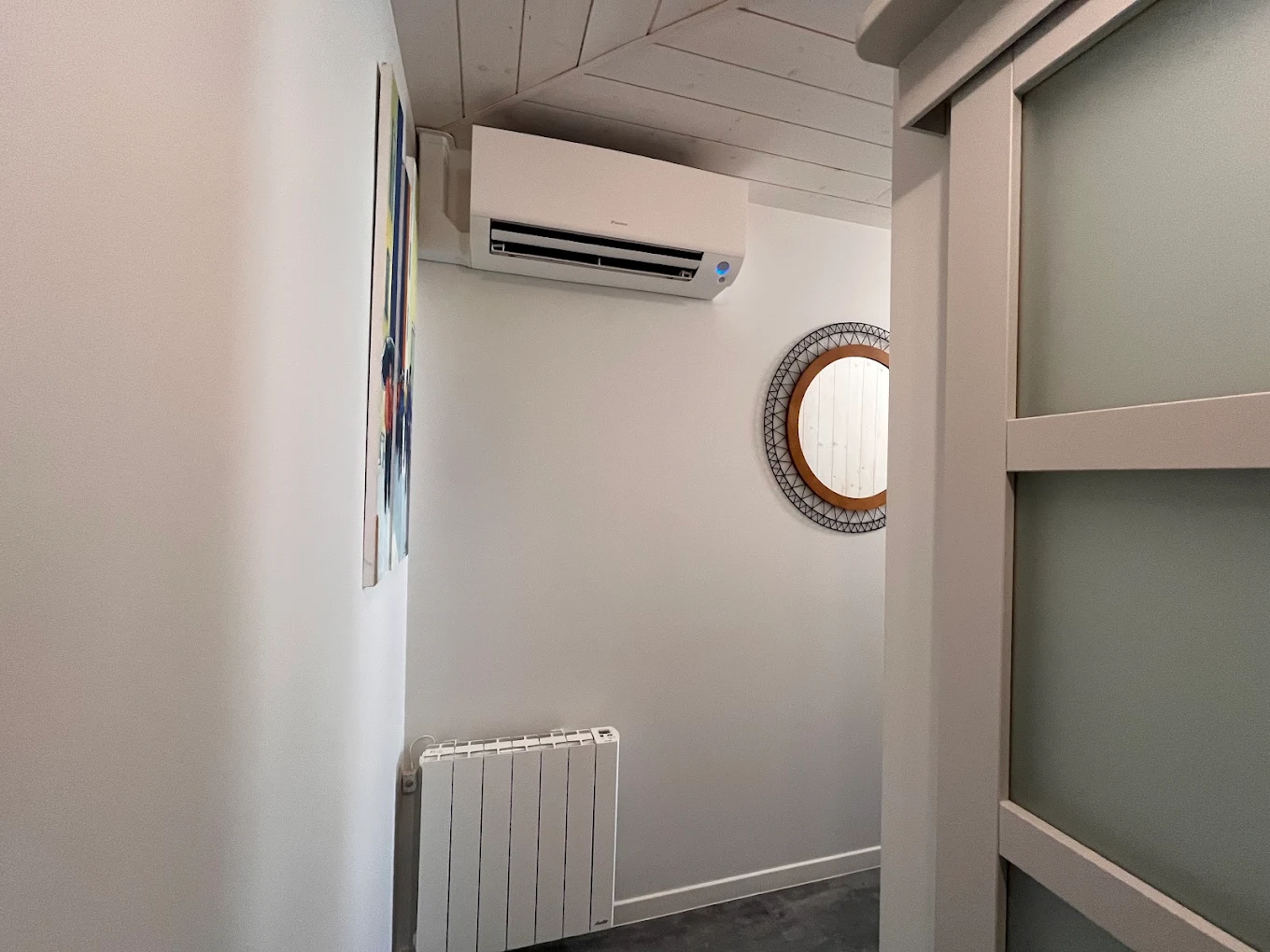 Unité intérieure de climatisation posée sous un plafond lambrissé blanc, à côté d'un radiateur existant